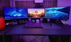 Ein episches **gaming set up** mit drei Monitoren, beleuchteter Tastatur und Maus. Im Hintergrund leuchtet lila Beleuchtung.